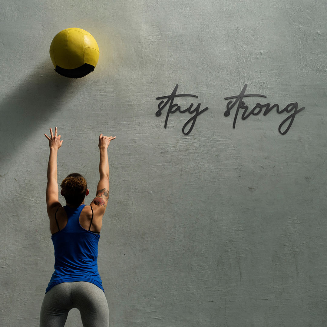 Wanddekoration Mesi Stay Strong an einer rauen Wand während eine sehr sportliche Frau einen Ball hoch wirft